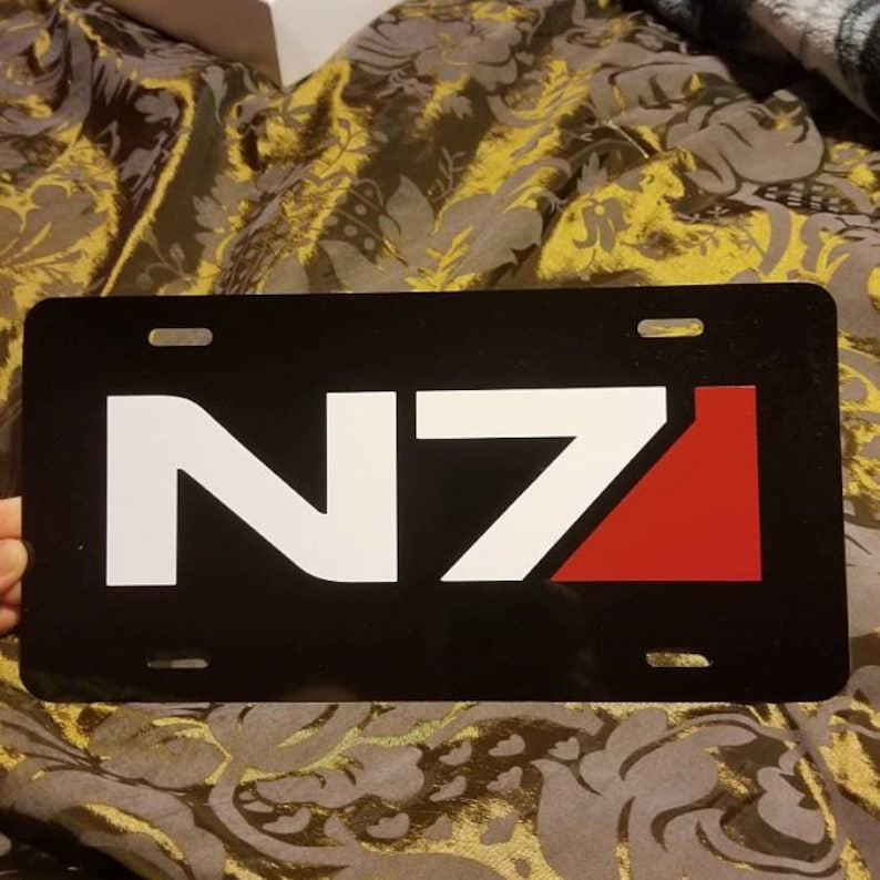 May include: Black license plate featuring a white N7 logo and a red triangle. The logo is a popular symbol from the Mass Effect video game series.