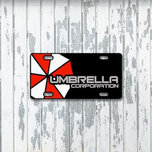Puede incluir: Placa de matrícula negra con el logotipo de Umbrella Corporation. El logotipo es un diseño de paraguas rojo y blanco. El texto "UMBRELLA CORPORATION" es blanco. La placa tiene cuatro orificios de montaje.