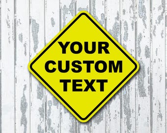 Puede incluir: Un letrero amarillo en forma de diamante con un borde negro y las palabras "YOUR CUSTOM TEXT" en letras negras en negrita. El letrero está sobre un fondo de madera blanca desgastada.