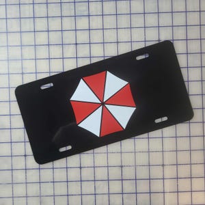 Op de afbeelding: Zwarte nummerplaat met een rood-wit paraplu-logo. De plaat heeft vier montagegaten en een glanzende afwerking. Het paraplu-ontwerp is gecentreerd op de plaat.