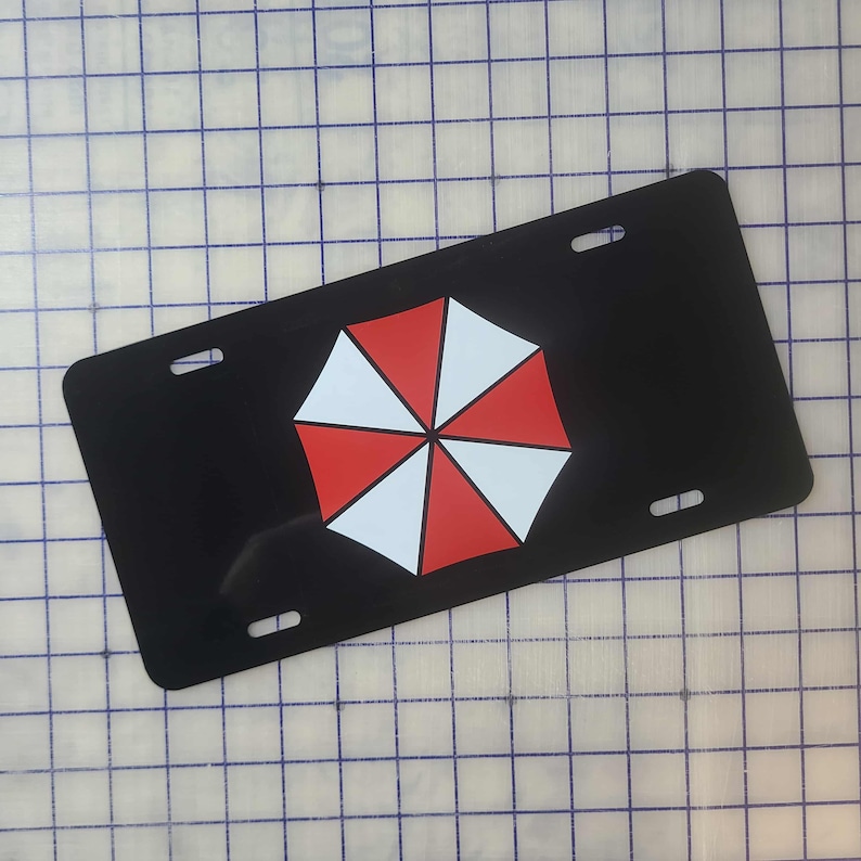 Op de afbeelding: Zwarte nummerplaat met een rood-wit paraplu-logo. De plaat heeft vier montagegaten en een glanzende afwerking. Het paraplu-ontwerp is gecentreerd op de plaat.