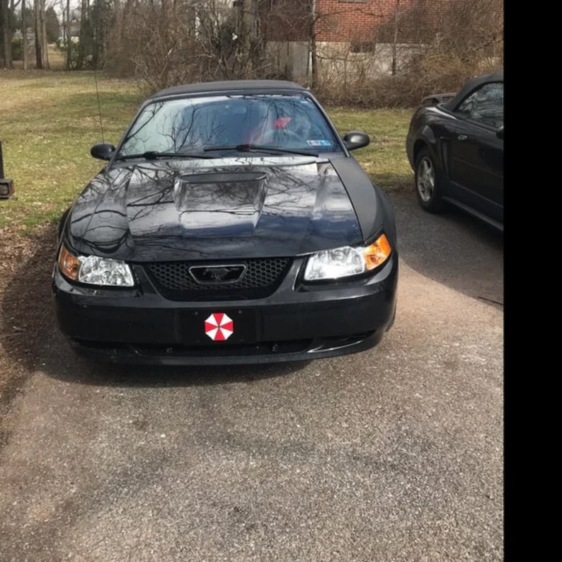Op de afbeelding: Een zwarte Ford Mustang cabriolet met een motorkapschep en een voorplaat met een rood-wit paraplu-logo. De auto staat geparkeerd op een verharde oprit, met een andere zwarte cabriolet zichtbaar op de achtergrond.