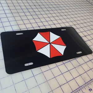 Op de afbeelding: Zwarte nummerplaat met een rood, wit en zwart paraplu-logo. De plaat heeft vier montagegaten en een glanzende afwerking. Het paraplu-ontwerp is een gestileerd, gesegmenteerd patroon.