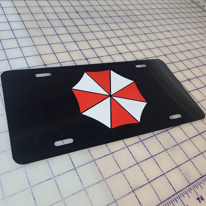 Op de afbeelding: Zwarte nummerplaat met een rood, wit en zwart paraplu-logo. De plaat heeft vier montagegaten en een glanzende afwerking. Het paraplu-ontwerp is een gestileerd, gesegmenteerd patroon.