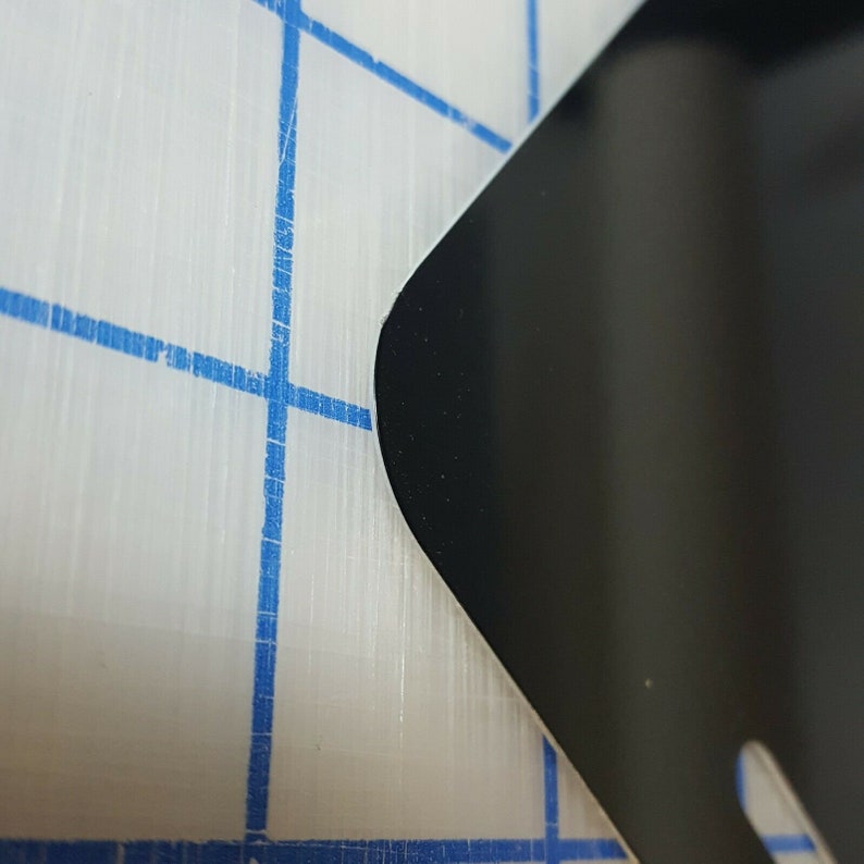 Op de afbeelding: Close-up van een zwart, gebogen object met een afgeronde rand. Het object staat tegen een wit oppervlak met een blauw rasterpatroon, wat een contrast in texturen en kleuren cre&euml;ert. De afbeelding benadrukt het gladde, donkere oppervlak.