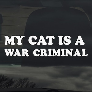 May include: White text on a dark background reads "MY CAT IS A WAR CRIMINAL." The text is in a bold, sans-serif font. The image appears to be a sticker or decal, possibly for a car window.