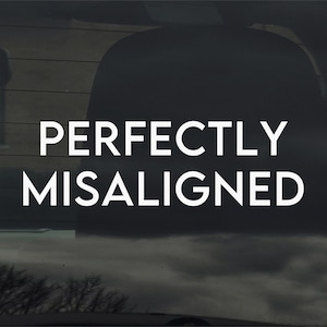 以下が含まれることがあります： 白い太字で「PERFECTLY MISALIGNED」と書かれた車の窓。背景には、曇り空と木のぼやけた景色があり、テキストと対照的です。