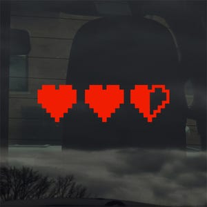May include: A red pixelated heart sticker showing three hearts, one partially filled, on a car window. The design is reminiscent of a video game health bar, with a retro aesthetic.
