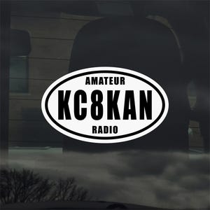 Op de afbeelding: Witte ovale sticker met zwarte tekst die "AMATEUR KC8KAN RADIO" luidt. De sticker is waarschijnlijk voor een autoruit of een vergelijkbaar oppervlak. De achtergrond is een vage weergave van een auto-interieur en de buitenomgeving.