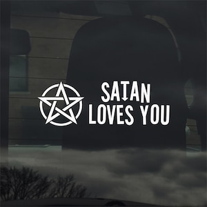 Puede incluir: Calcomanía blanca con un pentagrama dentro de un círculo y el texto "SATAN LOVES YOU" en negrita, sin serifa. Diseñada para su aplicación en una ventana de coche o superficie similar.