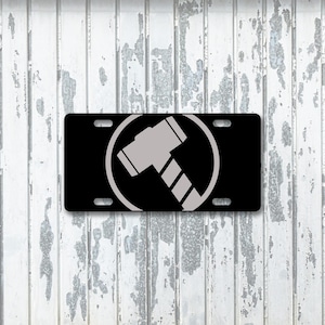 May include: Black license plate featuring a gray hammer design within a circle. The hammer has a detailed handle and head. The plate has four mounting holes and is set against a weathered, white wooden background.