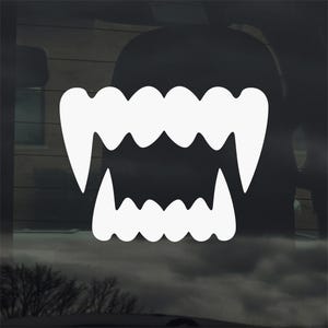 May include: A white vinyl decal featuring a stylized set of vampire fangs. The design includes upper and lower rows of teeth with sharp points and a jagged edge. The decal is suitable for application on various surfaces.