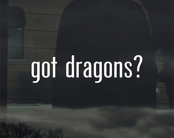 May include: A car window sticker with the text "got dragons?" in white. The background is dark, with a blurred view of the outside visible through the glass. The text is in a clean, sans-serif font.