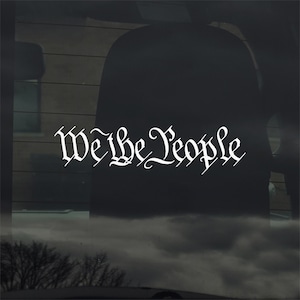 Pode incluir: Autocolante de vinil branco com as palavras "We the People" numa fonte decorativa. O autocolante é exibido numa janela, com um interior escuro e um céu nublado visível através do vidro. O texto está numa escrita estilizada.