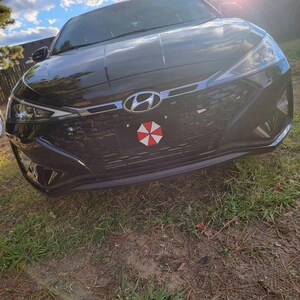 Op de afbeelding: Een zwarte Hyundai sedan met een kentekenplaat aan de voorkant met een rood, wit en zwart paraplu-logo. De auto staat op gras onder een gedeeltelijk bewolkte hemel. Het Hyundai-embleem is zichtbaar aan de voorkant.