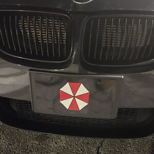 Op de afbeelding: Zwarte nummerplaat met een rood, wit en zwart paraplu-logo, gemonteerd op de voorkant van een zwarte auto. De grille van de auto is zichtbaar boven de nummerplaat.