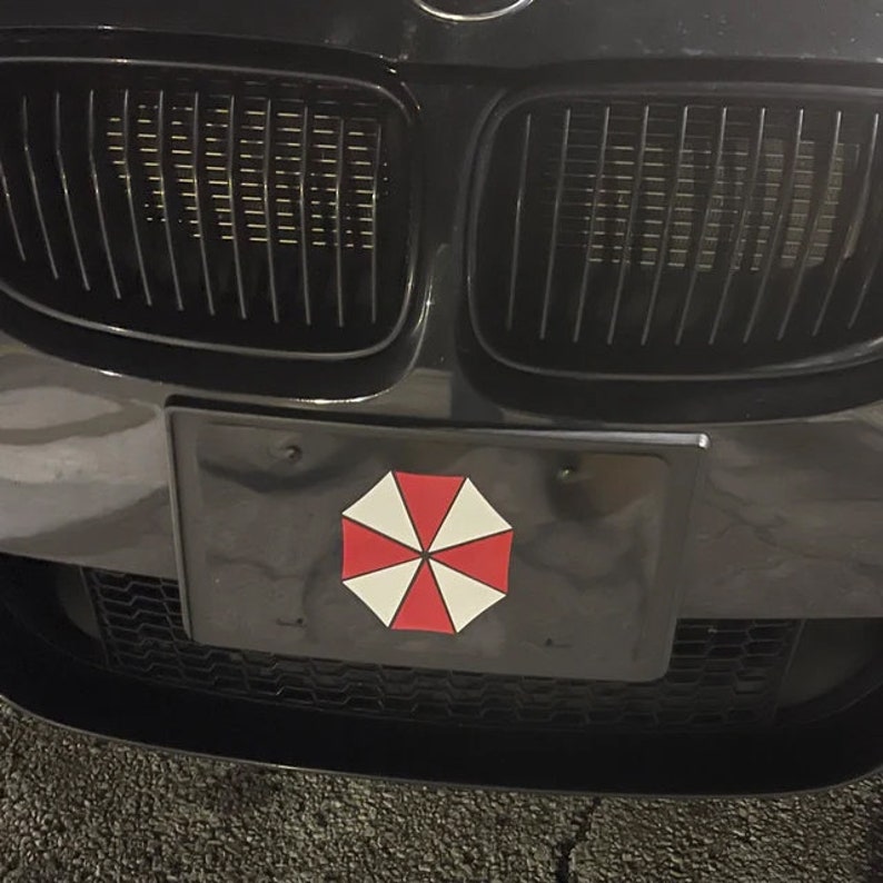Op de afbeelding: Zwarte nummerplaat met een rood, wit en zwart paraplu-logo, gemonteerd op de voorkant van een zwarte auto. De grille van de auto is zichtbaar boven de nummerplaat.
