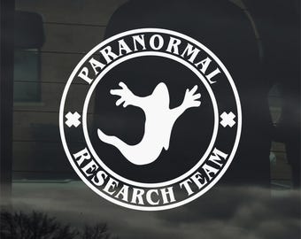 Könnte beinhalten: Weißer Vinyl-Aufkleber mit den Worten "PARANORMAL RESEARCH TEAM" in kreisförmigem Muster. Eine weiße Geistergrafik befindet sich in der Mitte. Der Aufkleber ist für die Anbringung an einer Autoscheibe oder ähnlichen Oberflächen konzipiert.
