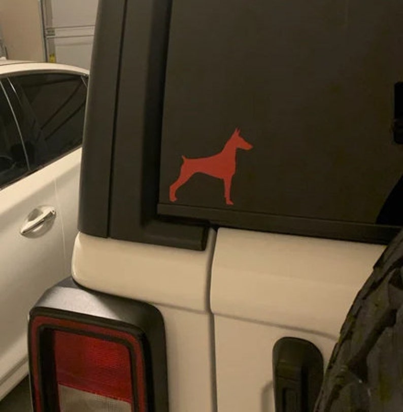 May include: A white car with a red silhouette of a dog on the back window. The car has a black tire and a black rear bumper.