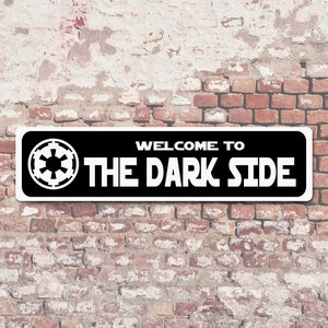 以下が含まれることがあります： 「WELCOME TO THE DARK SIDE」の文字と白いインペリアルシンボルが描かれた、白黒の長方形の看板。風化したレンガの壁に取り付けられています。
