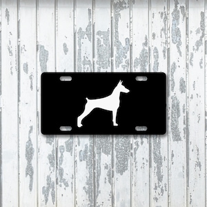 Puede incluir: Placa de matrícula negra con una silueta blanca de un perro Doberman Pinscher. La placa tiene cuatro orificios de montaje y un acabado brillante. El perro se representa en una pose de pie, con su cuerpo de perfil.