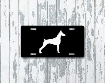 May include: Black license plate featuring a white silhouette of a Doberman Pinscher dog. The plate has four mounting holes and a glossy finish. The dog is depicted in a standing pose, with its body in profile.