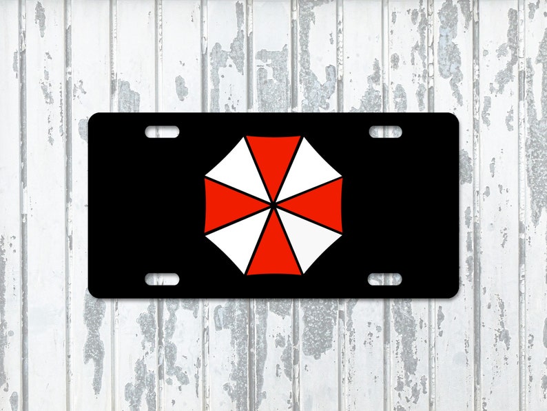 Op de afbeelding: Zwarte nummerplaat met een rood-wit paraplu-logo. Het logo staat in het midden van de plaat, die vier montagegaten heeft. Het ontwerp is een gedurfde afbeelding op een rechthoekige plaat.