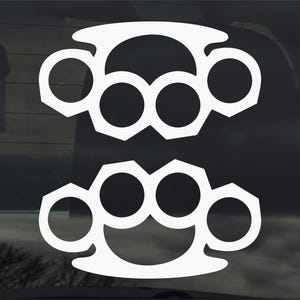 May include: White vinyl sticker of two brass knuckles, a popular symbol of strength and power. The sticker is designed to be applied to a car window or other smooth surface.