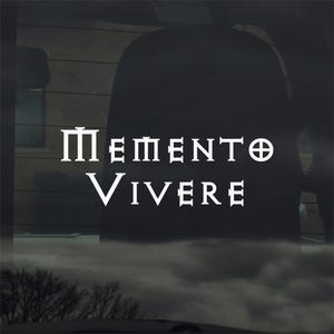 May include: White vinyl decal with the Latin phrase "Memento Vivere" in a bold, serif font. The decal is designed to be applied to a car window or similar surface. The background is a blurred view of the outside.