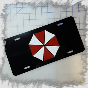 Op de afbeelding: Zwarte nummerplaat met een rood en wit paraplu-logo. Het logo is een segmentontwerp. De plaat heeft vier montagegaten en een glanzende afwerking. Een decoratief item voor fans van een bepaalde franchise.