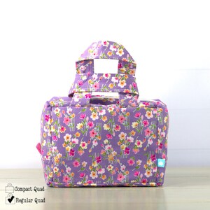 May include: A floral print tote bag in shades of purple, pink, and yellow. The bag features a rectangular shape with a handle and a top flap. The text "Compact Quad" and "Regular Quad" are visible on the bottom left.