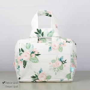 May include: A white fabric lunch bag featuring a repeating pattern of soft pink roses and green leaves. The bag has two top handles, a rectangular base, and a zippered closure. The design includes horizontal stitching lines across the surface.