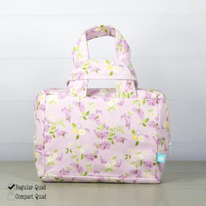 May include: A rectangular fabric bag featuring a light purple background with a repeating floral pattern of small purple and white flowers and green leaves. The bag has two top handles and a side zipper closure with a small blue tag attached.
