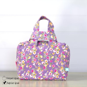 May include: A floral print tote bag in a lavender hue, adorned with pink, yellow, and white flowers. The bag features a rectangular shape, a top handle, and a small side pocket. The design includes the text "Regular Quad".