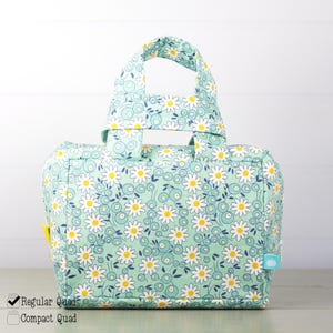 May include: A rectangular fabric bag with two top handles, featuring a light mint green background patterned with white daisies with yellow centers and small blue berries. A small blue tag is visible on the side with a white pumpkin icon.