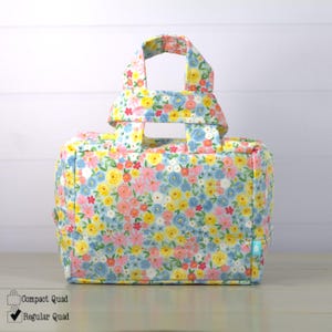 May include: A floral print tote bag with a rectangular shape and a top handle. The bag features a pattern of pink, yellow, and blue flowers on a white background. The bag is designed with a compact quad and regular quad option.