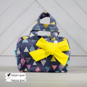 May include: A small, blue fabric handbag with a yellow bow and a geometric pattern of triangles. The bag has a top handle and a zipper closure. The text "Compact Quad" and "Regular Quad" is visible in the image.