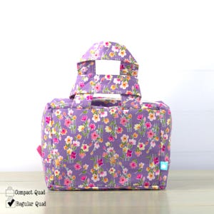 May include: A floral print tote bag in shades of purple, pink, and yellow. The bag has a rectangular shape with a handle and a top flap. The text "Compact Quad" and "Regular Quad" are visible on the bottom left.
