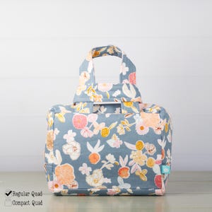 Peut inclure: Un sac en tissu rectangulaire avec deux poignées supérieures, présentant un fond bleu avec un motif floral répétitif dans des tons de rose, orange, jaune et blanc. Le sac a une forme structurée et une petite étiquette bleu clair sur le côté.