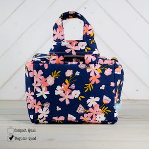 May include: A navy blue tote bag with a floral pattern in pink, white, and yellow. The bag has a rectangular shape with a handle on top. The floral design is repeated across the entire surface of the bag.