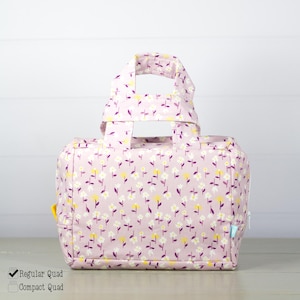 May include: A rectangular fabric bag with two top handles, featuring a light purple background with a repeating pattern of small white and yellow flowers. The bag has a zipper closure and a small light blue tag on the side. Text reads Regular Quad and Compact Quad.