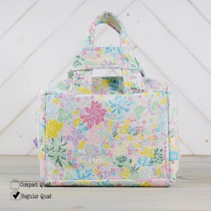 May include: A white canvas tote bag with a floral print featuring succulents in shades of pink, green, yellow, and blue. The bag has two handles and a rectangular shape.