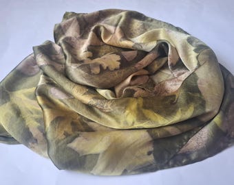 Può includere: Sciarpa di seta con stampa botanica. La sciarpa presenta un motivo di foglie nelle tonalità del verde, marrone e giallo. Il tessuto è drappeggiato, mostrando la consistenza e il movimento del materiale. È un accessorio da indossare.