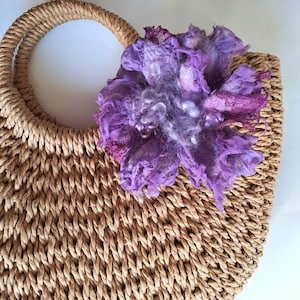 May include: A woven straw handbag with a rounded shape and a sturdy handle. A large, handmade purple felt flower is attached to the bag, adding a decorative touch. The flower has a textured appearance with shades of purple and white.