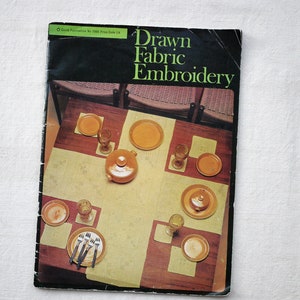 May include: A book titled "Drawn Fabric Embroidery" with a photograph of a table setting featuring yellow plates and glasses on a white tablecloth.