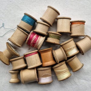 May include: A collection of vintage wooden spools filled with colourful threads. The spools are various sizes and hold threads in shades of blue, pink, orange, green, and cream. The spools are arranged on a light-coloured surface.