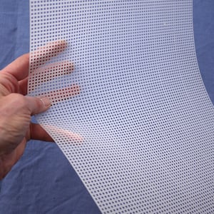 May include: A sheet of white plastic canvas with a grid of square holes. The canvas is held up by a hand, showing its flexibility. The background is a solid blue fabric.