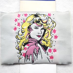 May include: White fabric patch with a pink and yellow embroidered image of a woman with long blonde hair. The background is white with pink stars.