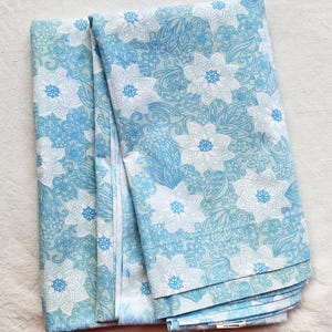 May include: A folded piece of fabric featuring a repeating pattern of white and light blue lotus-style flowers, smaller blossoms, and swirling leaf motifs set against a soft, pale blue background. The textile has a smooth, consistent texture.