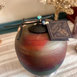 May include: A Raku Dream Jar with a round, bulbous shape. The jar has a dark, mottled finish with shades of red, green, and gold. A black lid is topped with a decorative leather piece and turquoise and blue beads. A tag reads "Raku Dream Jar."